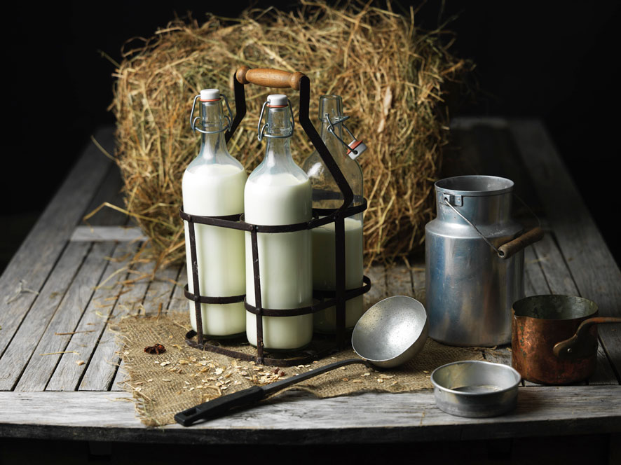 1 pot de lait en fer et 2 bouteilles en verre contenant du lait sont posés sur une table en bois. En arrière plan de la paille et devant une louche.Image illustrant le lait français collecté par Le Gall pour la fabrication de ses beurres et crèmes.