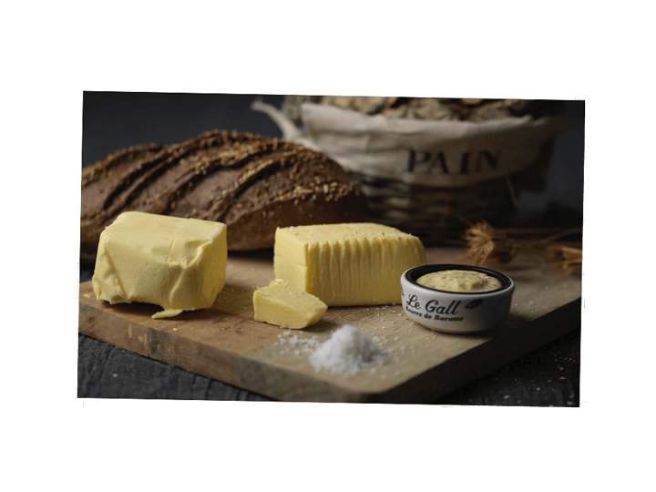 Tout savoir sur le beurre : beurre de baratte coupé en 2 sur une planche de bois avec un petit amas de sel. En arrière plan deux pains de campagne dans un dans sa panière rustique